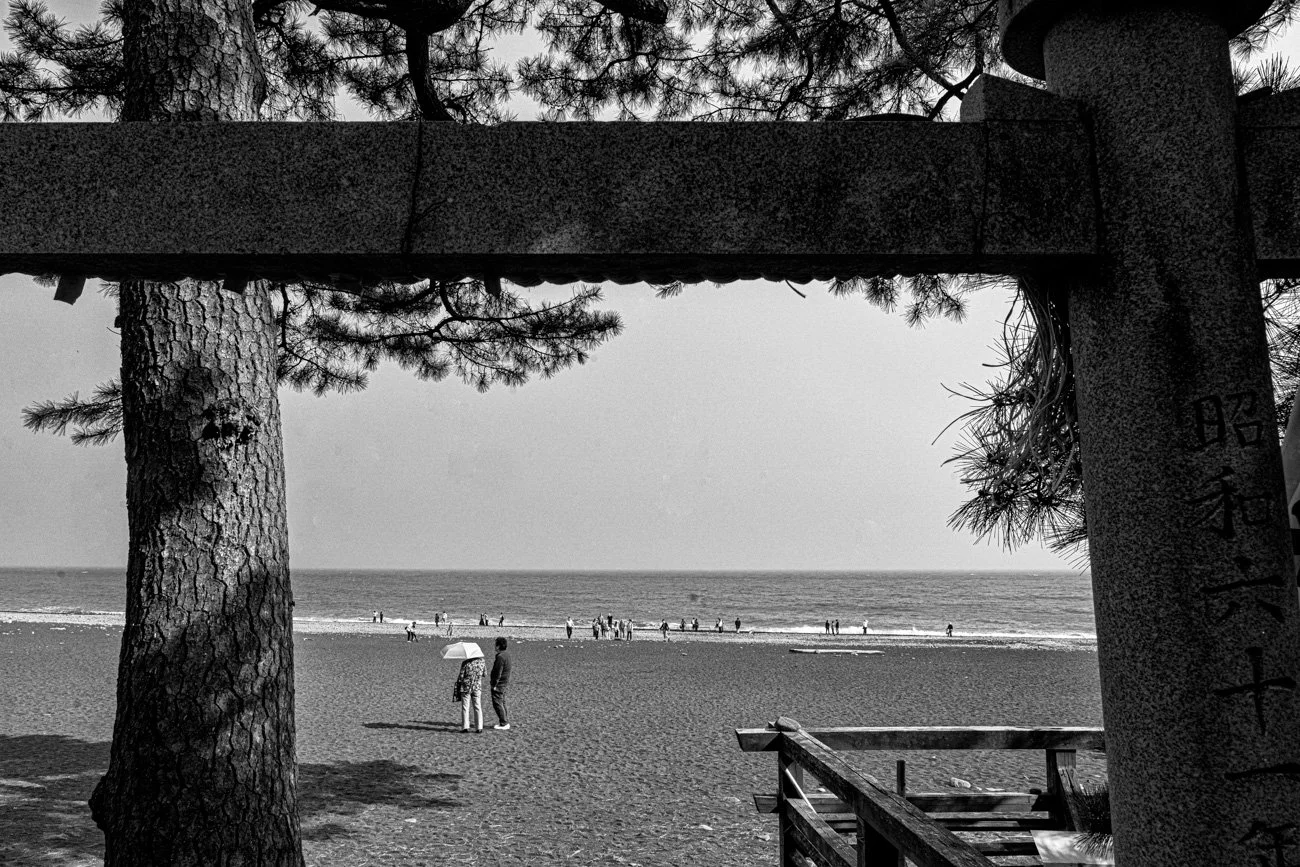 Black and white photo of a beach scene, framed by a stone torii gate, with people walking along the shoreline and trees in the background.