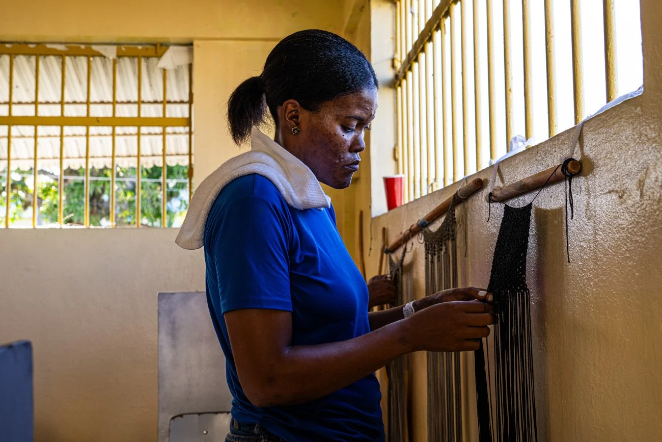 A woman weaving black beads on a wall loom in a well-lit room with barred windows.
