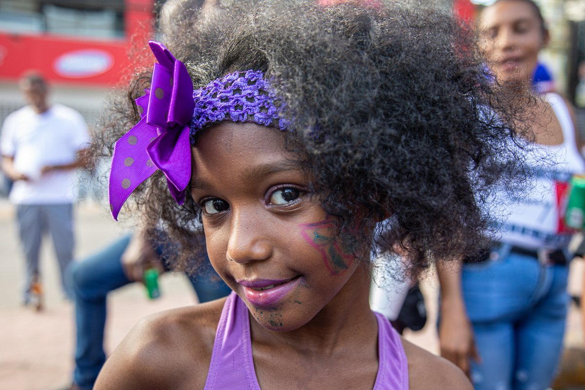 A young girl with curly hair, wearing a purple headband and tank top, has face paint on her cheek and mouth, and a large purple bow accessory in her hair. She is outside at a festive event.