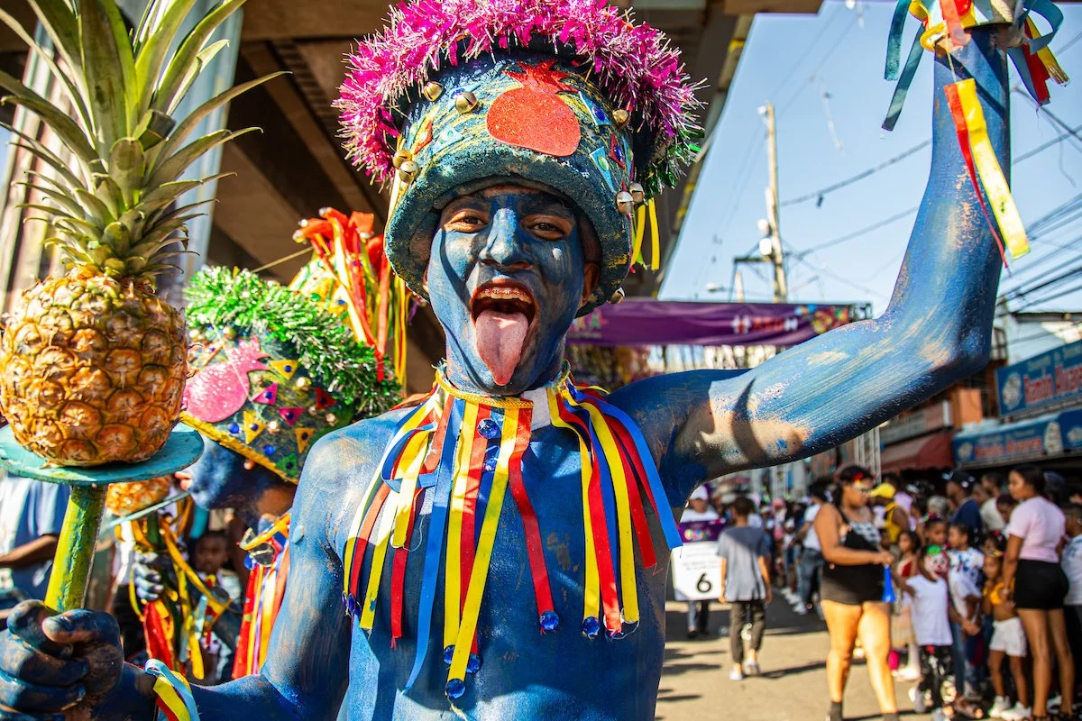 A person painted blue wearing a colorful costume with ribbons, a hat decorated with tinsel, and holding a pineapple on a stick during a street celebration or parade.