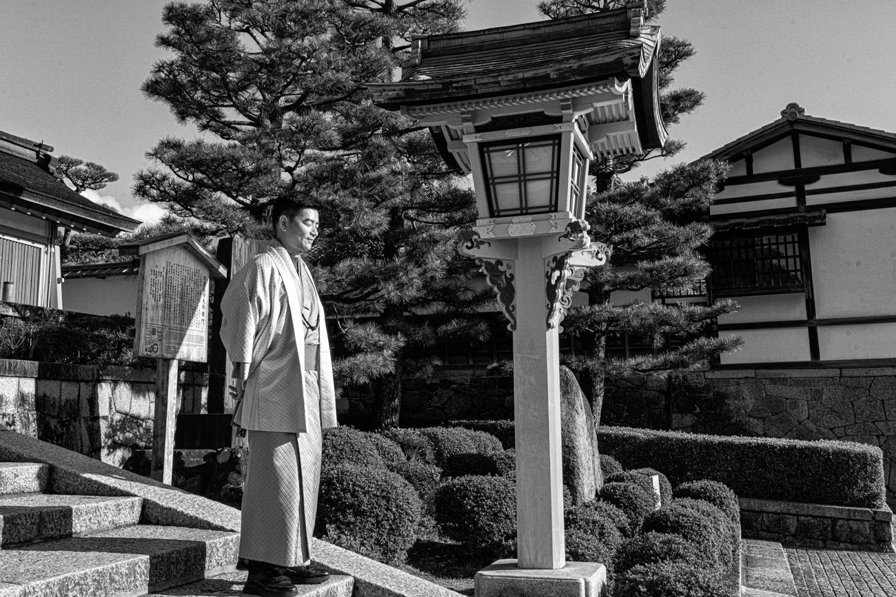 A man in traditional Japanese attire standing on stone steps outside a temple, with a large lamp post and manicured bushes, trees, and buildings in the background.
