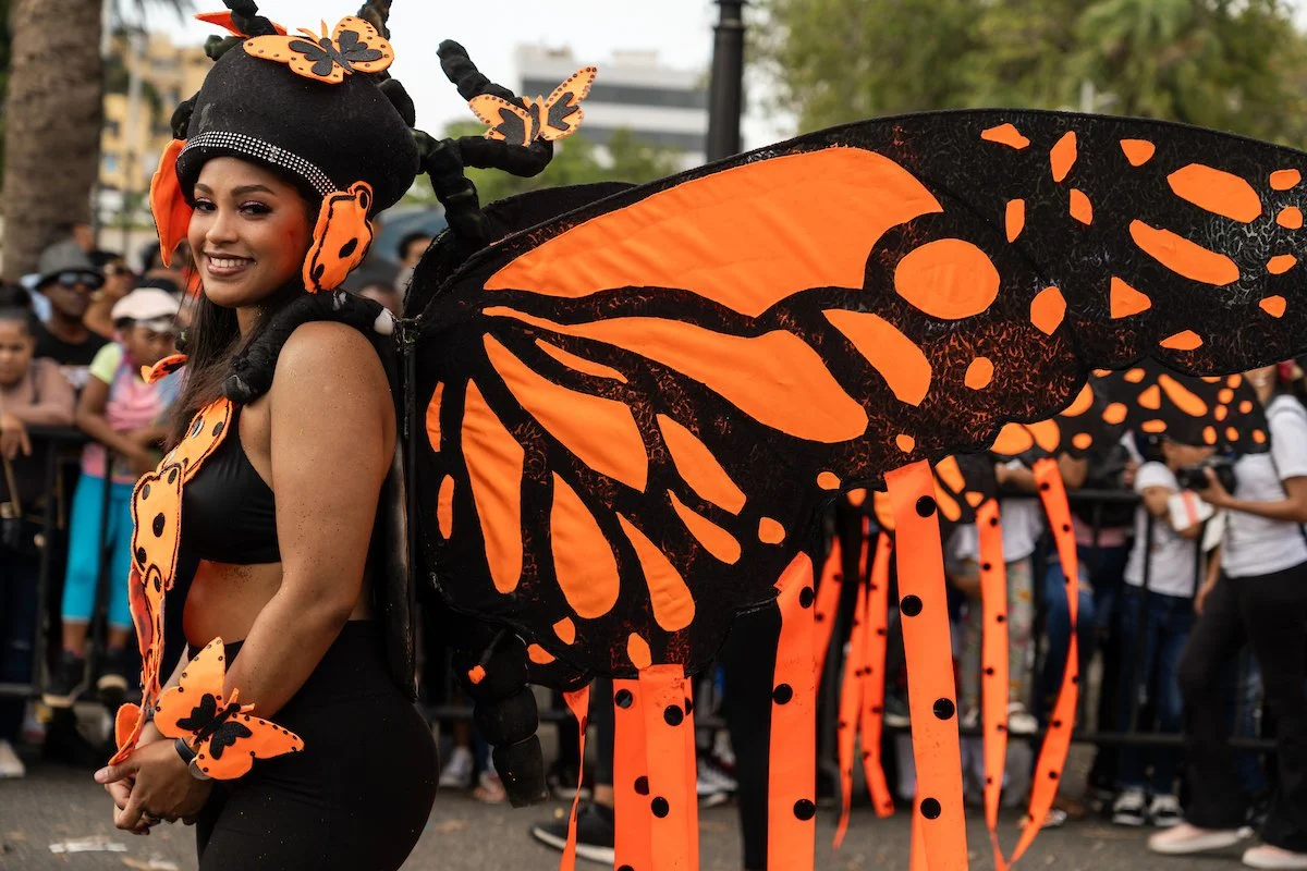 A woman in a butterfly costume with large orange and black wings, butterfly accessories, and a black outfit, at a parade with a crowd in the background.