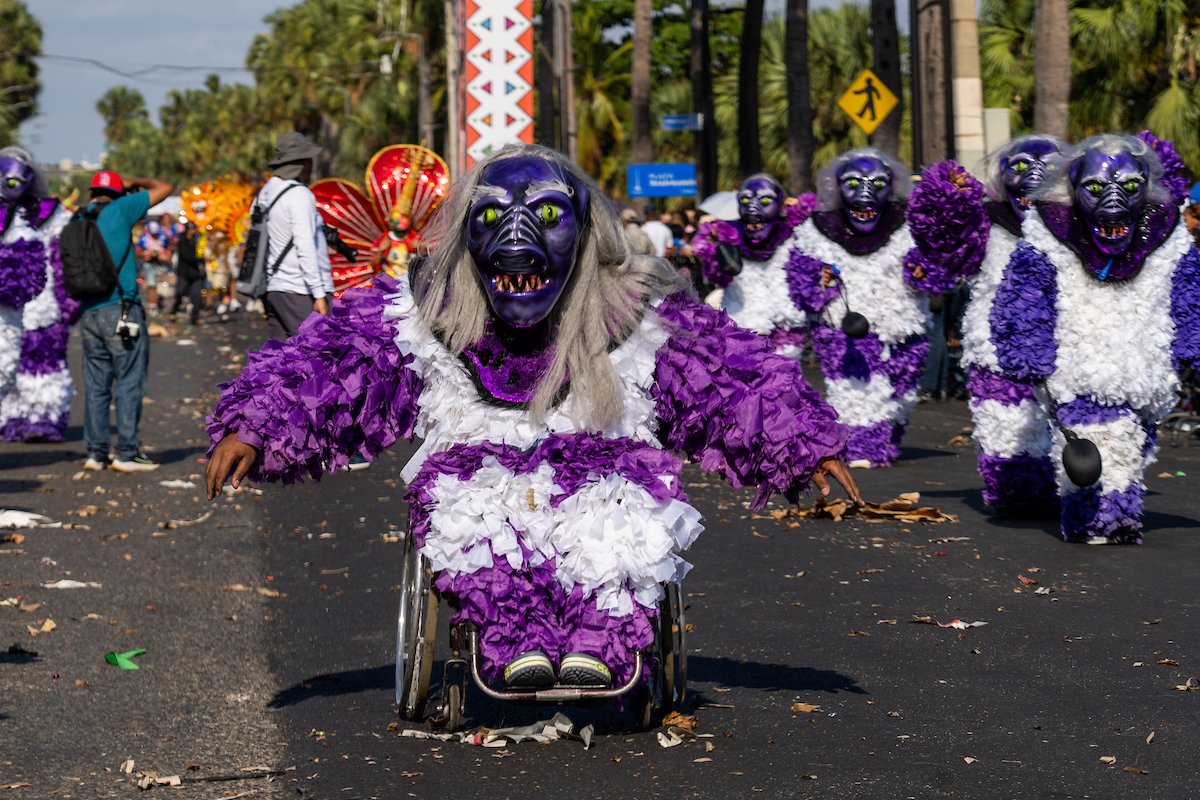 A person in a wheelchair dressed in a purple and white costume with a purple mask with green eyes and large teeth, participating in a parade with other similarly dressed parade participants in the background.