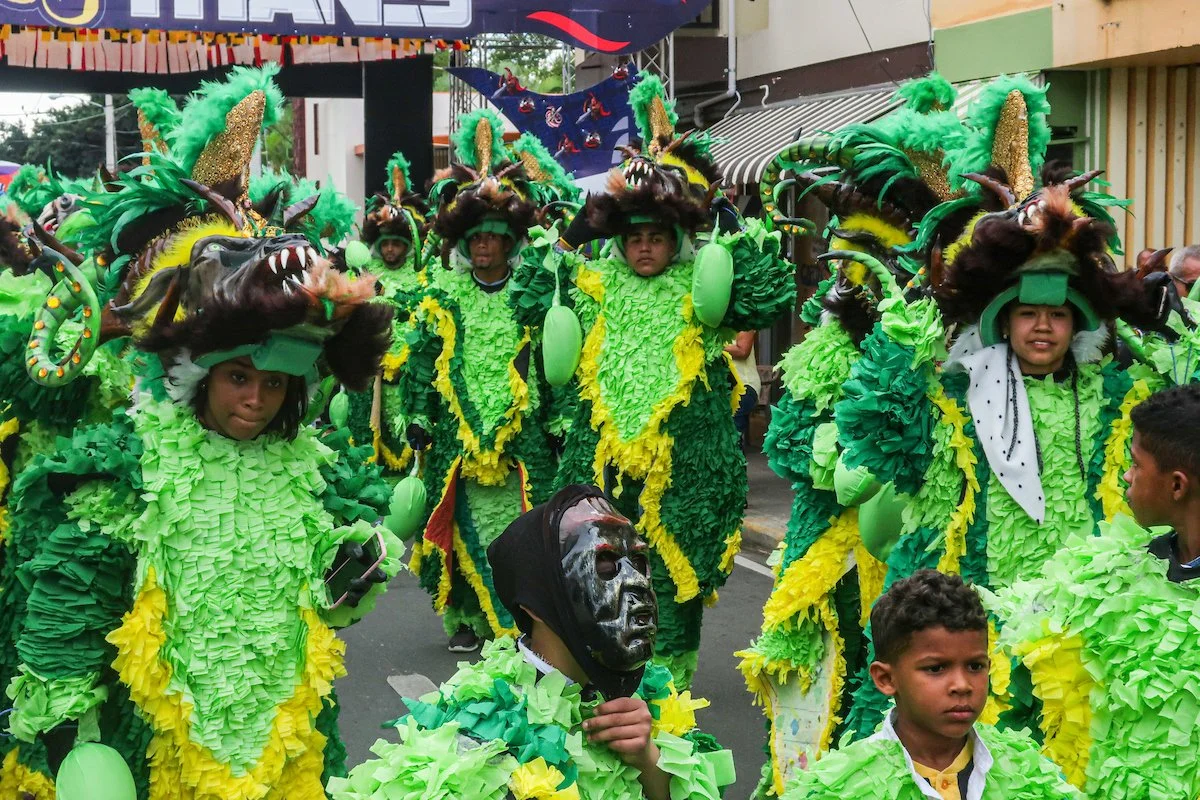 Children dressed in colorful, elaborate costumes with green, yellow, and black feathers and masks, participating in a parade or festival.
