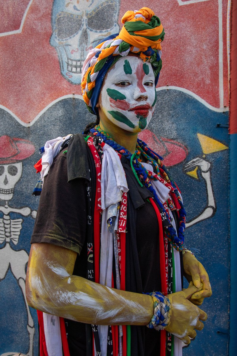 Person with painted face, wearing multicolored headwrap and multiple beaded necklaces, standing in front of a colorful mural featuring a skeleton and balloons.
