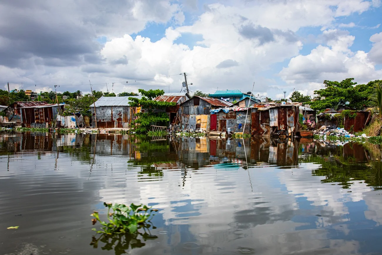 A shanty settlement by a body of water, with rusted metal houses and trash, under a cloudy sky.