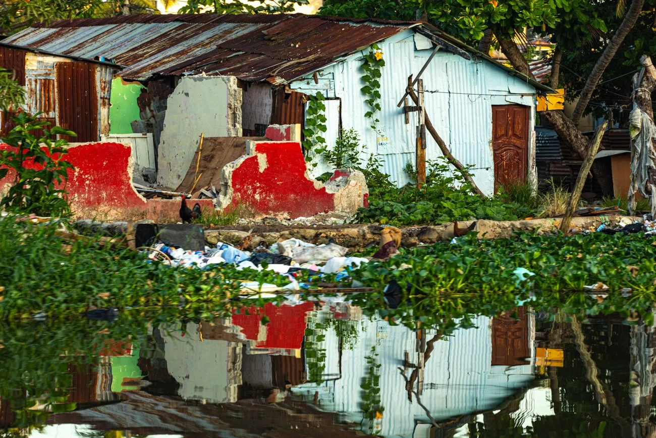A small, rundown house with a rusted metal roof, peeling wall paint, and surrounded by vegetation. There is garbage floating in the water in front of the house, indicating water pollution or flooding.
