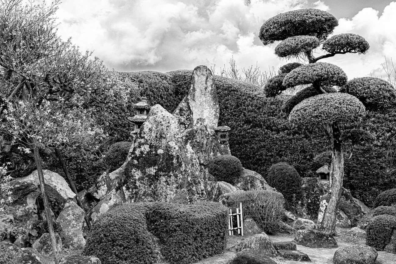 A Japanese-style garden with trimmed trees, large rocks, and stone lanterns, under a cloudy sky.