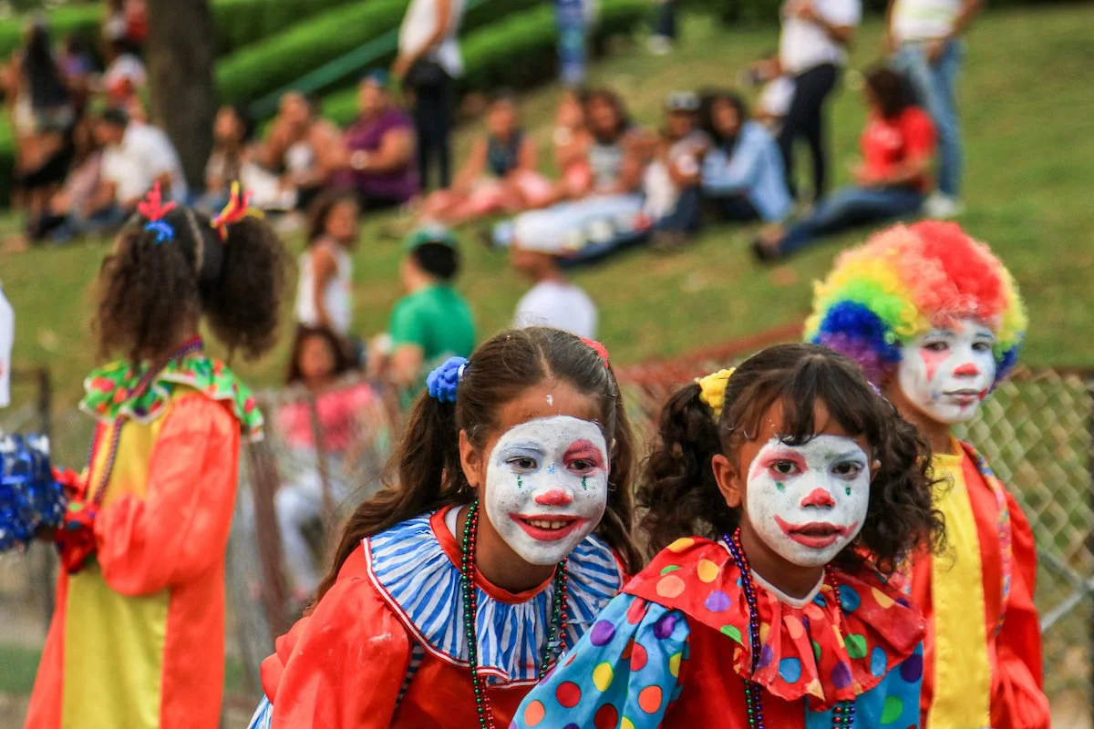 Three children dressed as clowns with painted faces and colorful costumes, standing outdoors at a gathering or festival, with a blurred crowd in the background.