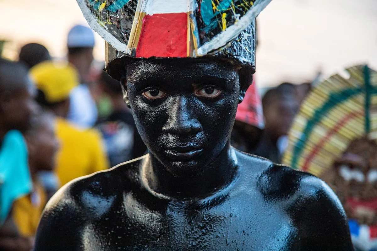 A person with black body paint and wearing a colorful headdress during a cultural event or festival, with a crowd in the background.