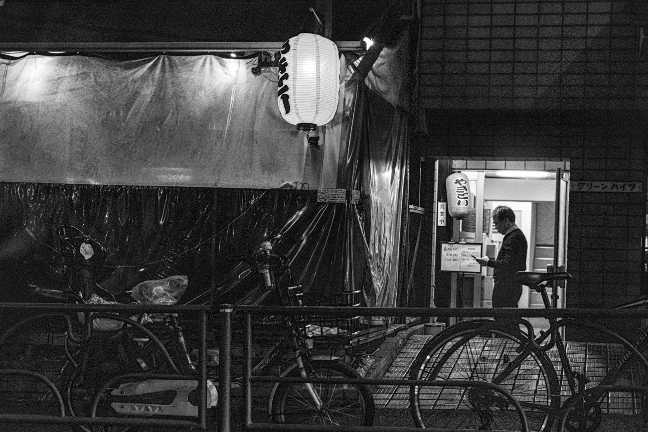 A man standing outside a small Japanese restaurant at night, using his phone, illuminated by exterior lights, with bicycles parked in front.