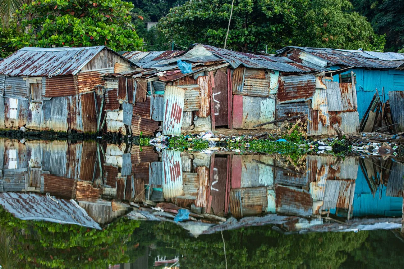 A reflection of a dilapidated shack made of corrugated metal sheets and wood in a body of water. Green foliage surrounds the shack.