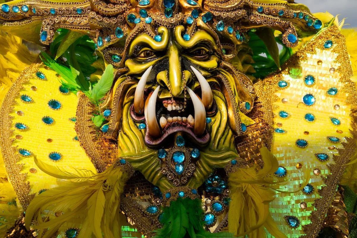 Colorful, elaborate costume featuring a fierce, ornate mask with exaggerated facial features, large tusks, and surrounded by yellow feathers, blue gemstones, and decorative fabric.