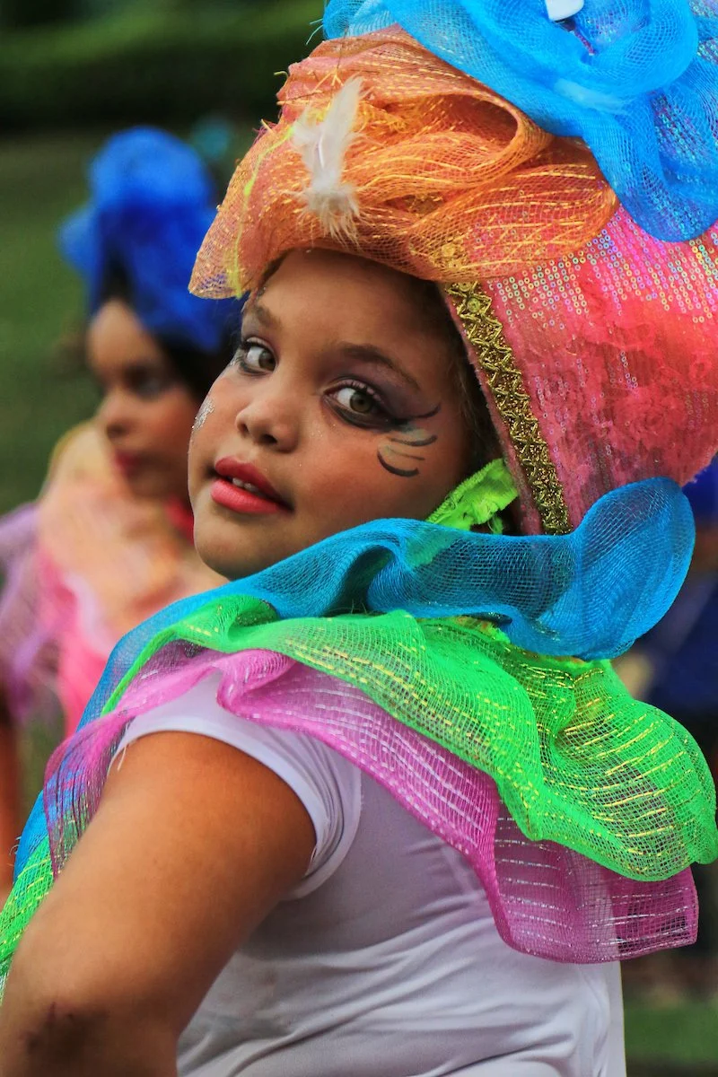 Two children dressed in colorful, glittery costumes with large, flowy, multicolored fabric headpieces. The girl in front has face paint and is looking at the camera, while the girl in the background is turned slightly to the side with her face partia