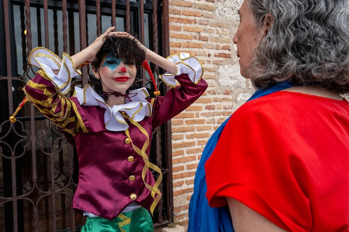 A person dressed as a clown with colorful makeup, a maroon and gold costume, and curly dark hair, standing outside in front of a brick wall and engaging with an older person wearing a red and blue shirt.