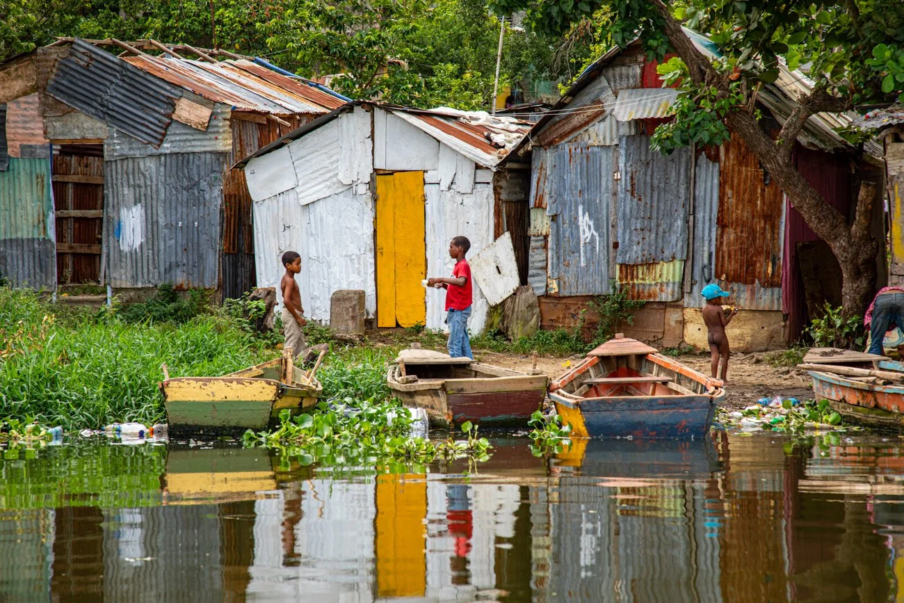 A group of children playing near the water in front of makeshift houses made of corrugated metal sheets, with boats floating on the water and surrounded by green vegetation.