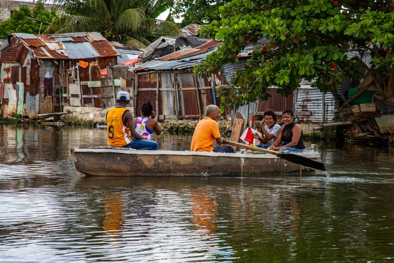 A group of five people in a small boat navigating a waterway in a neighborhood with makeshift houses made of rusted metal sheets and lush greenery overhead.