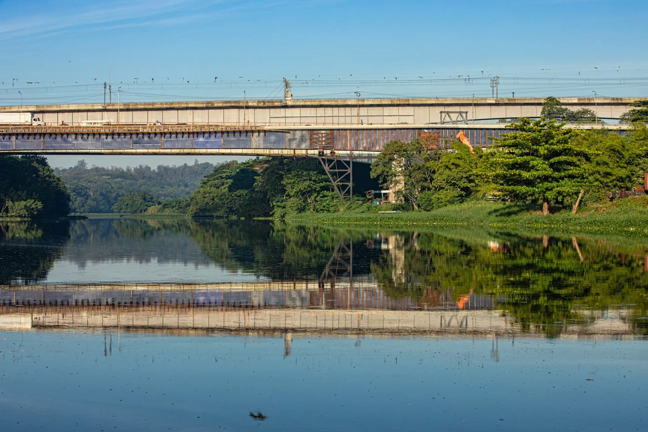 A body of water reflecting a bridge, trees, and the sky with few clouds.