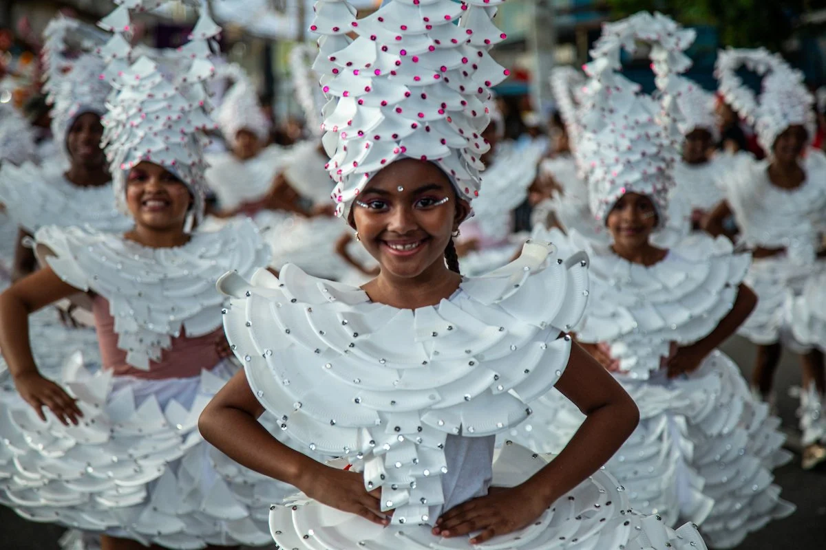 Young girls participating in a parade or celebration, dressed in elaborate white costumes with ruffled layers and tall, matching headdresses adorned with small beads or ornaments, smiling and posing for the camera.