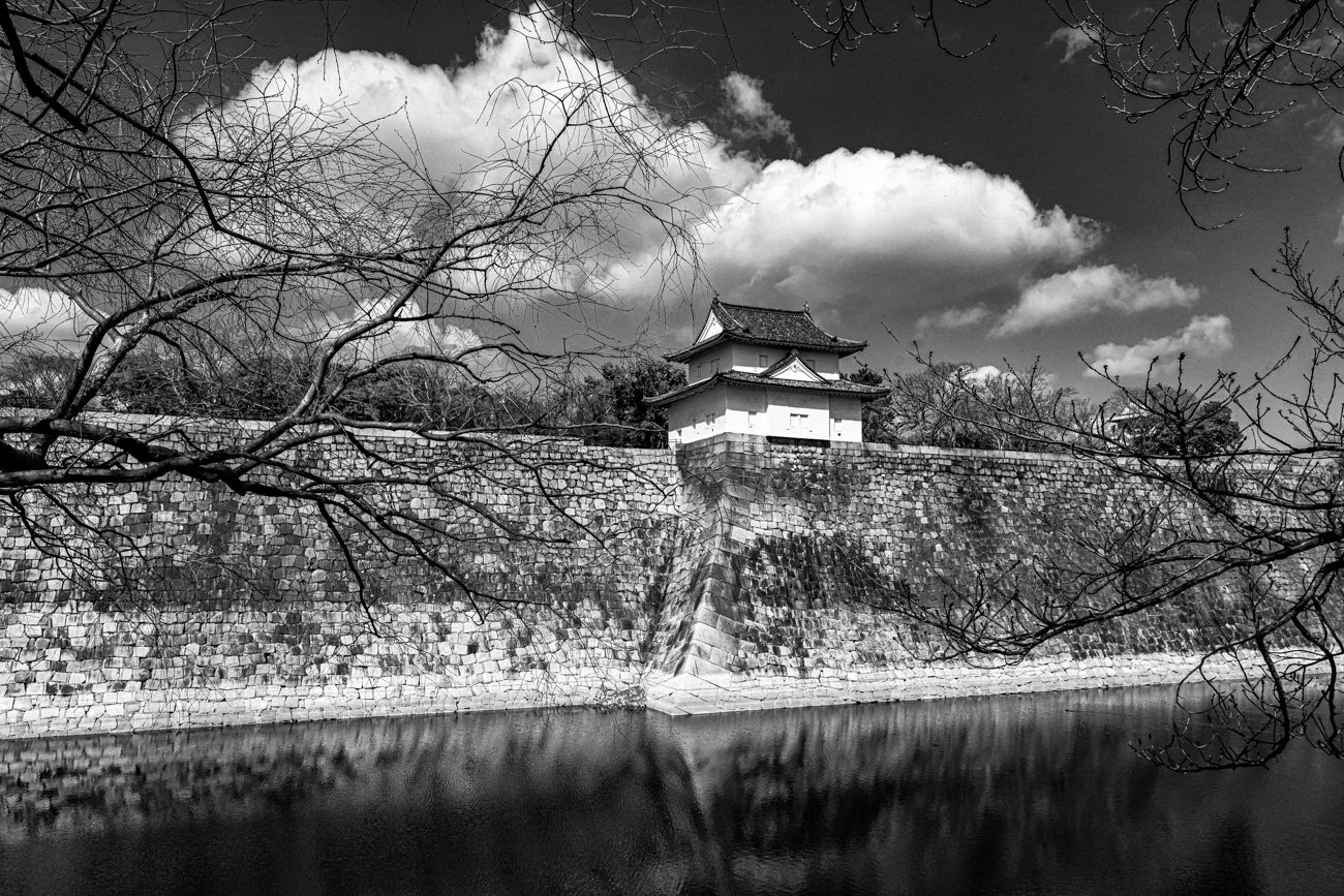 Black and white photo of a traditional Japanese castle sitting on a stone wall, with a moat in front and leafless trees nearby. The sky is partly cloudy.