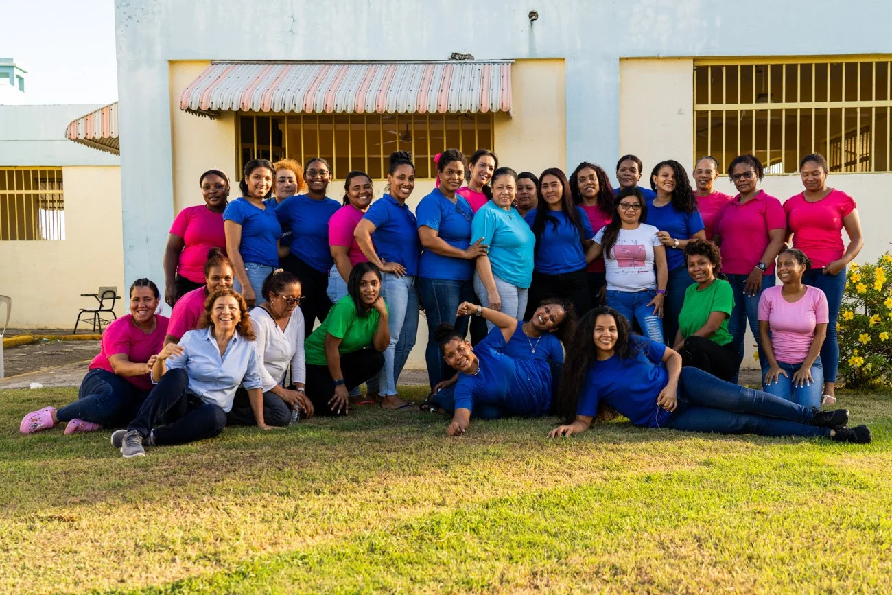 Group of women in colorful casual clothing posing outside on a grassy lawn in front of a building with yellow bars and awnings.