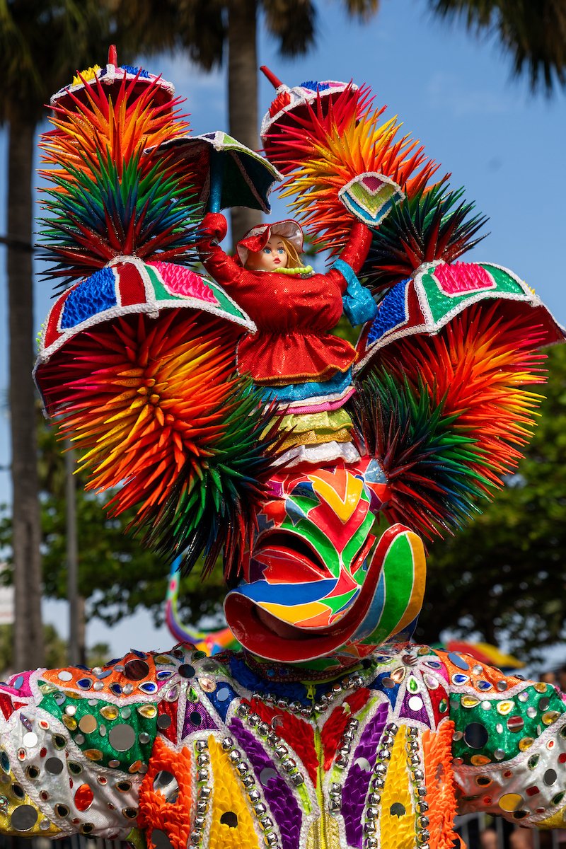 Colorful carnival costume with a mask and headdress made of spiky, multicolored, petal-like structures, and a doll-like face in the center.