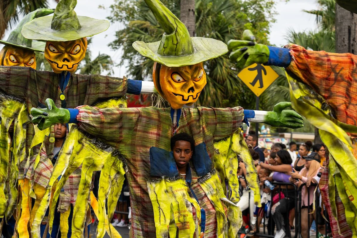 People participating in a parade wearing scarecrow costumes with yellow skull masks and wide-brimmed green hats, in front of a crowd of spectators