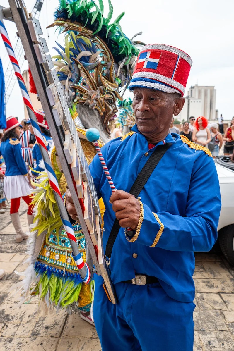 An elderly man dressed in a blue uniform with gold details and a red, white, and blue hat, holding a painted baton, standing near a person in an elaborate carnival costume with feathers and beads at a parade or festival.