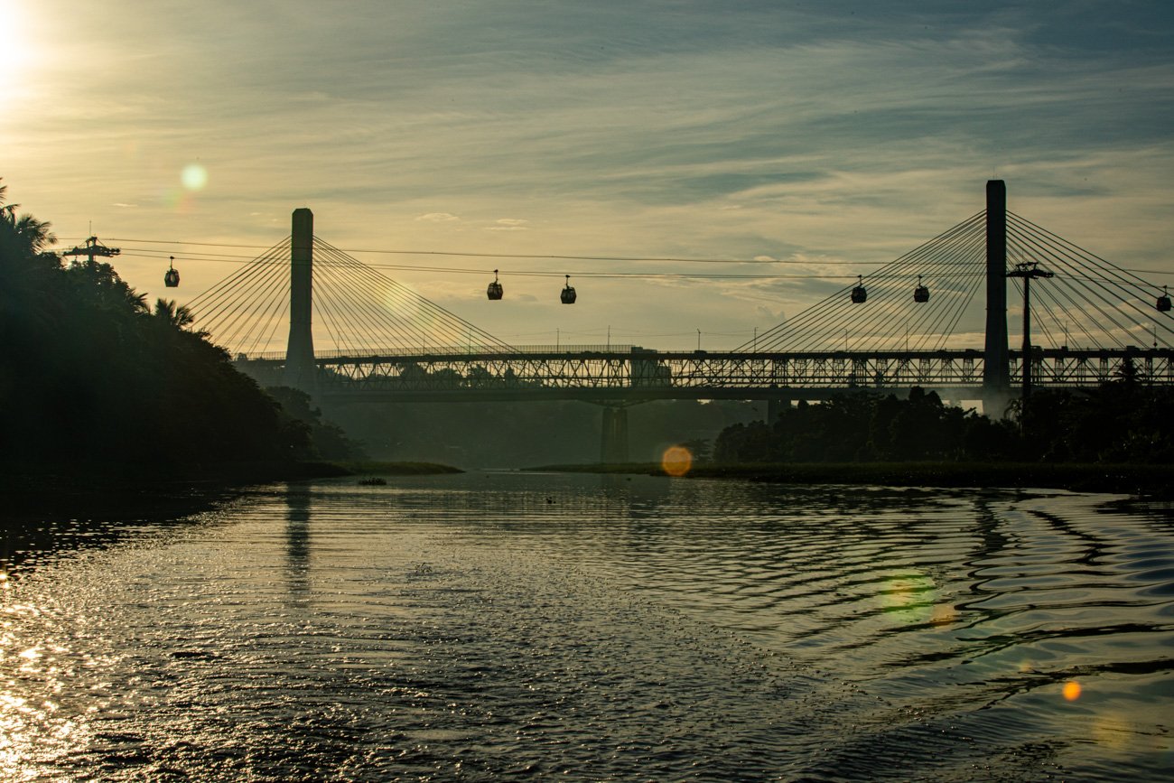 A photo of a river with a bridge and cable cars crossing above, during sunset with a partly cloudy sky.