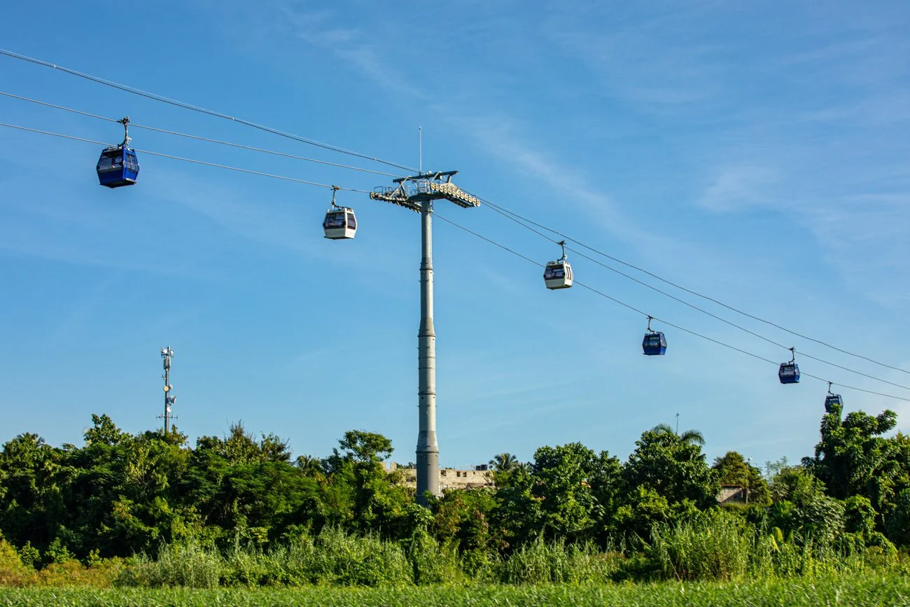 Cable cars suspended on cables over a lush green landscape with trees and a blue sky.