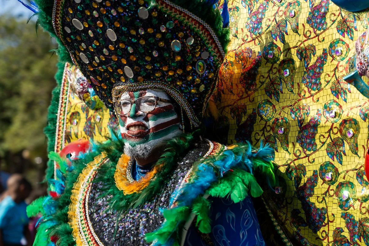 A man dressed in a colorful carnival costume with face paint in green, white, and red stripes, wearing glasses, and an elaborate headdress with sequins, feathers, and beaded accents. The costume is decorated with green feathers and a yellow collar, a