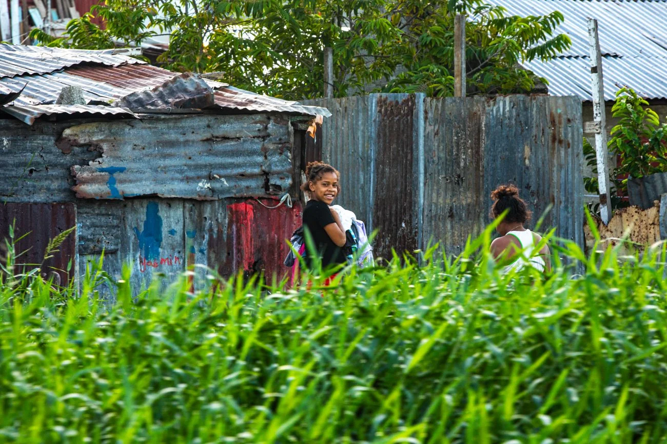 Two young girls smiling and playing outdoors near a rustic, corrugated metal fence, with tall green grass in the foreground.