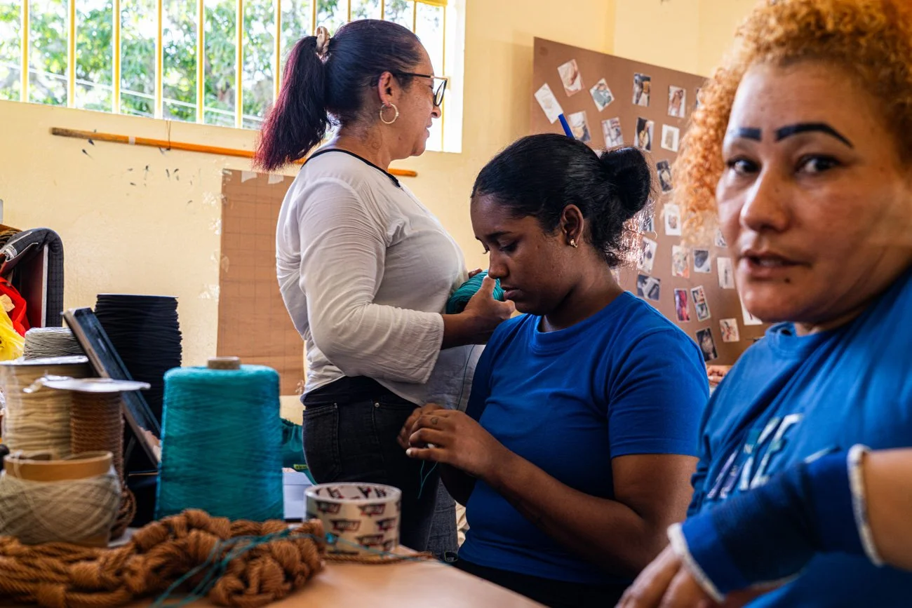 Three women working on a craft project with yarn and thread in a room with a bulletin board and a window.