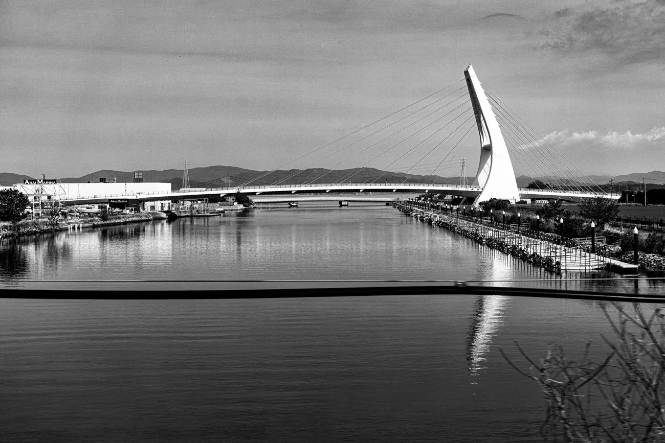 A black and white photo of a modern cable-stayed bridge with a large curved pylon, spanning over a calm river, with mountains and a partly cloudy sky in the background.