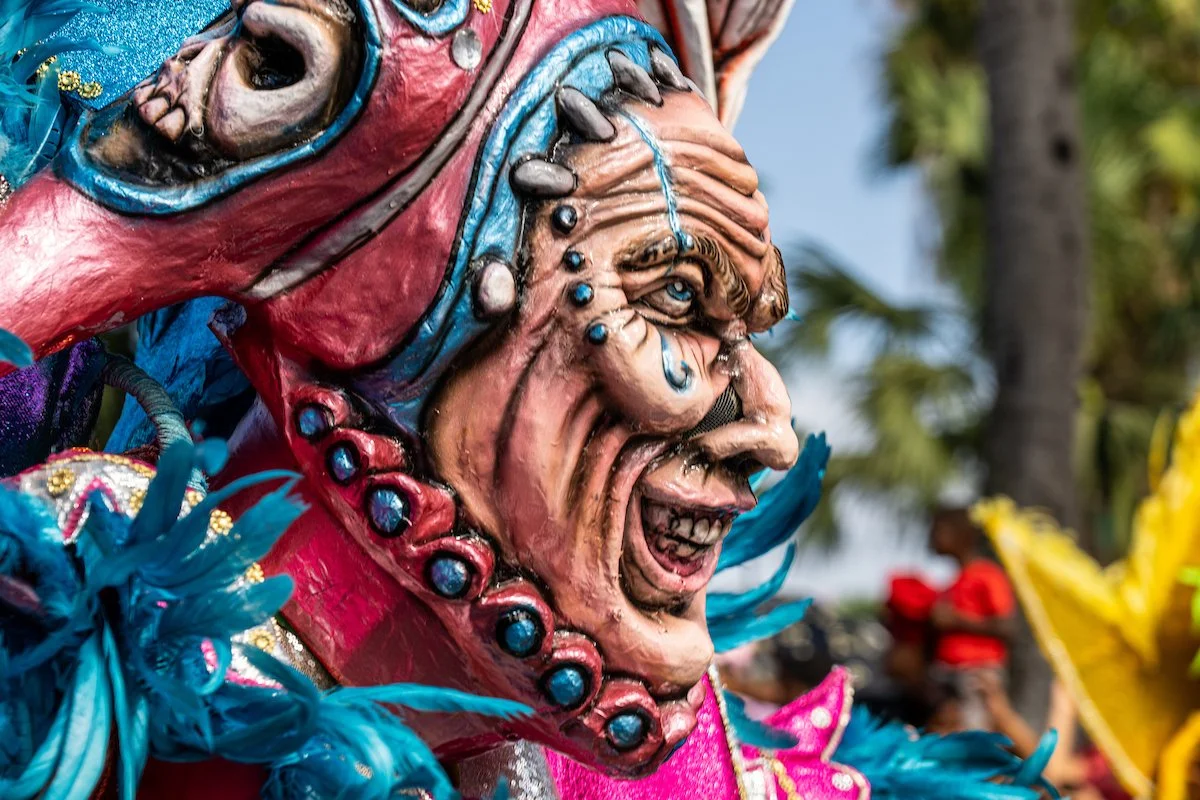 A colorful, elaborate costume mask depicting a smiling, wrinkled old man's face with blue eyes, decorated with beads, feathers, and vibrant fabrics, worn during a festive cultural celebration.