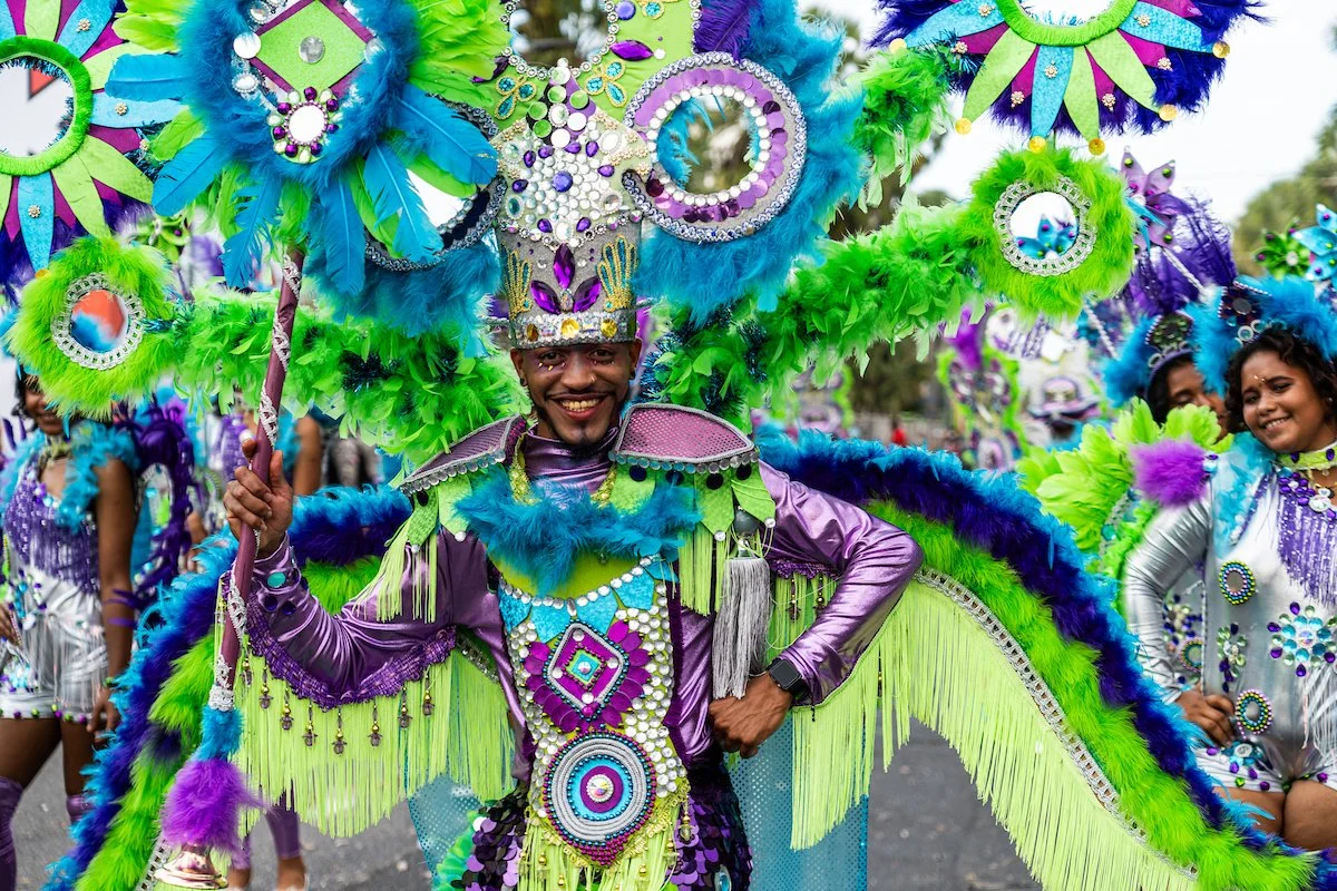 A man in colorful carnival costume smiling among other dancers in vibrant attire with large feathered and jeweled headdresses.