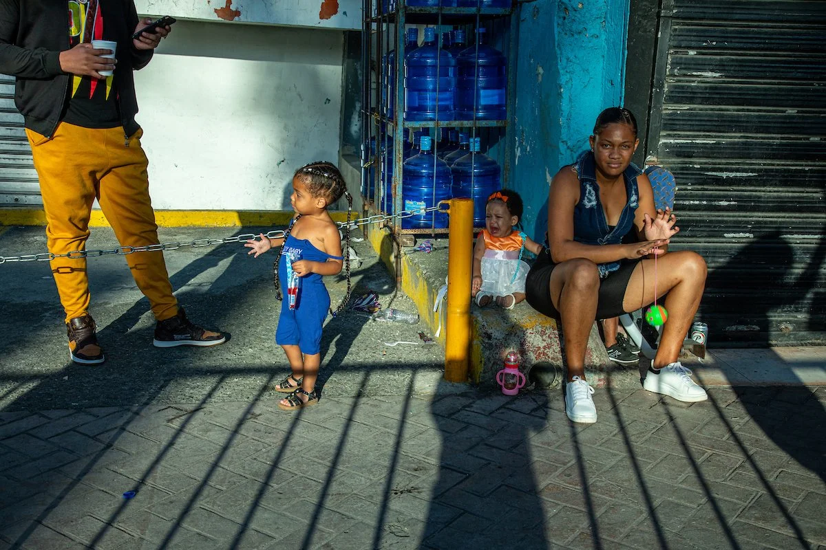 A woman sitting on a curb with two children next to her, one boy and one girl, on a street with water bottles in a cage behind them. The boy is standing and the girl is crying. The woman is looking at her phone. There are shadows on the ground and va