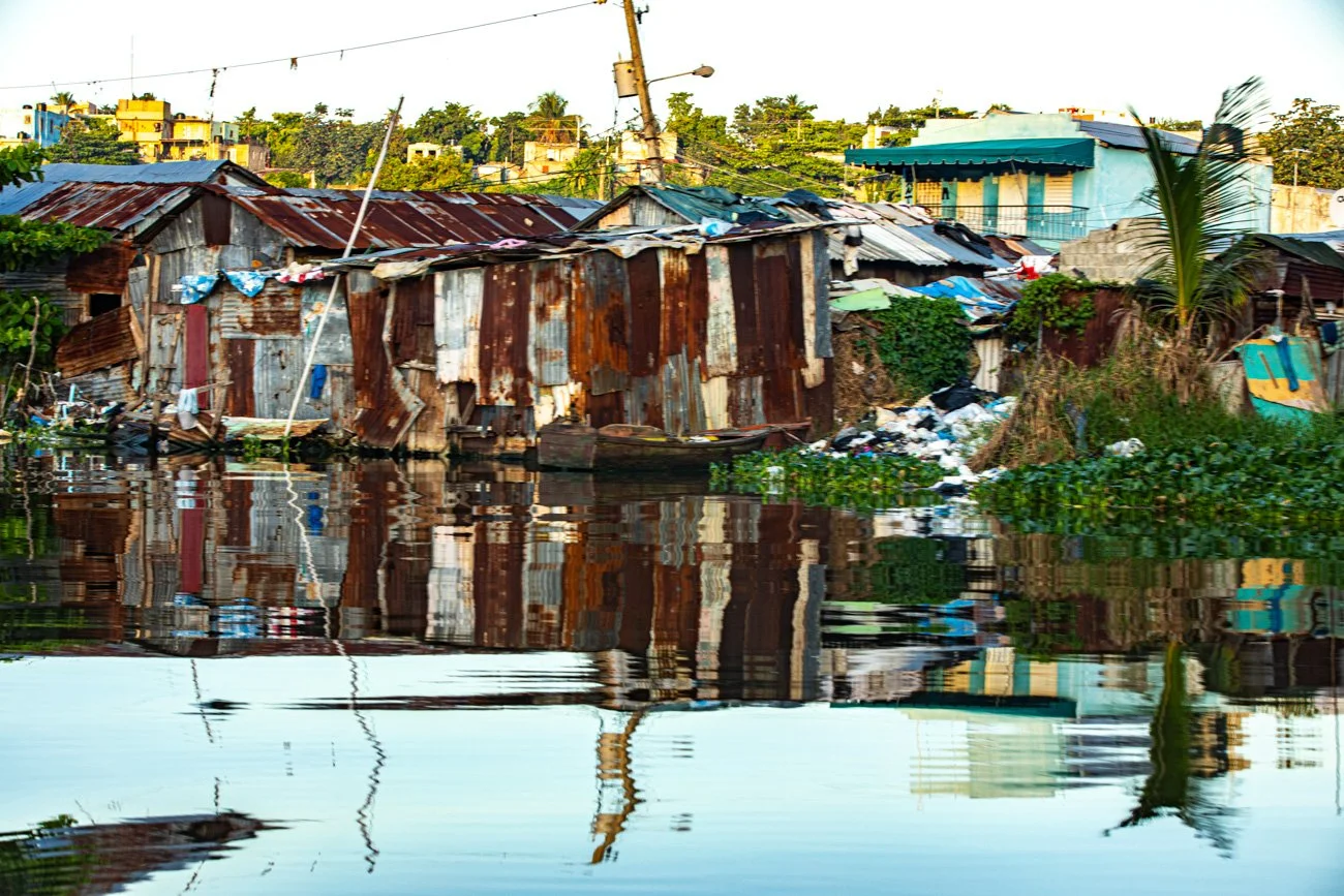 Rundown houses along a waterway with trash and vegetation, reflecting in the water, in an impoverished neighborhood.
