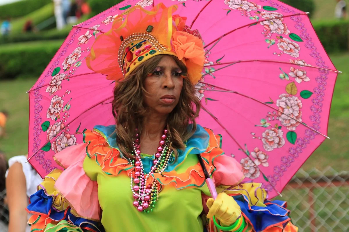 Woman dressed in colorful, ruffled attire holding a pink floral umbrella and wearing an elaborate headpiece and beaded necklaces at what appears to be a festive outdoor event.