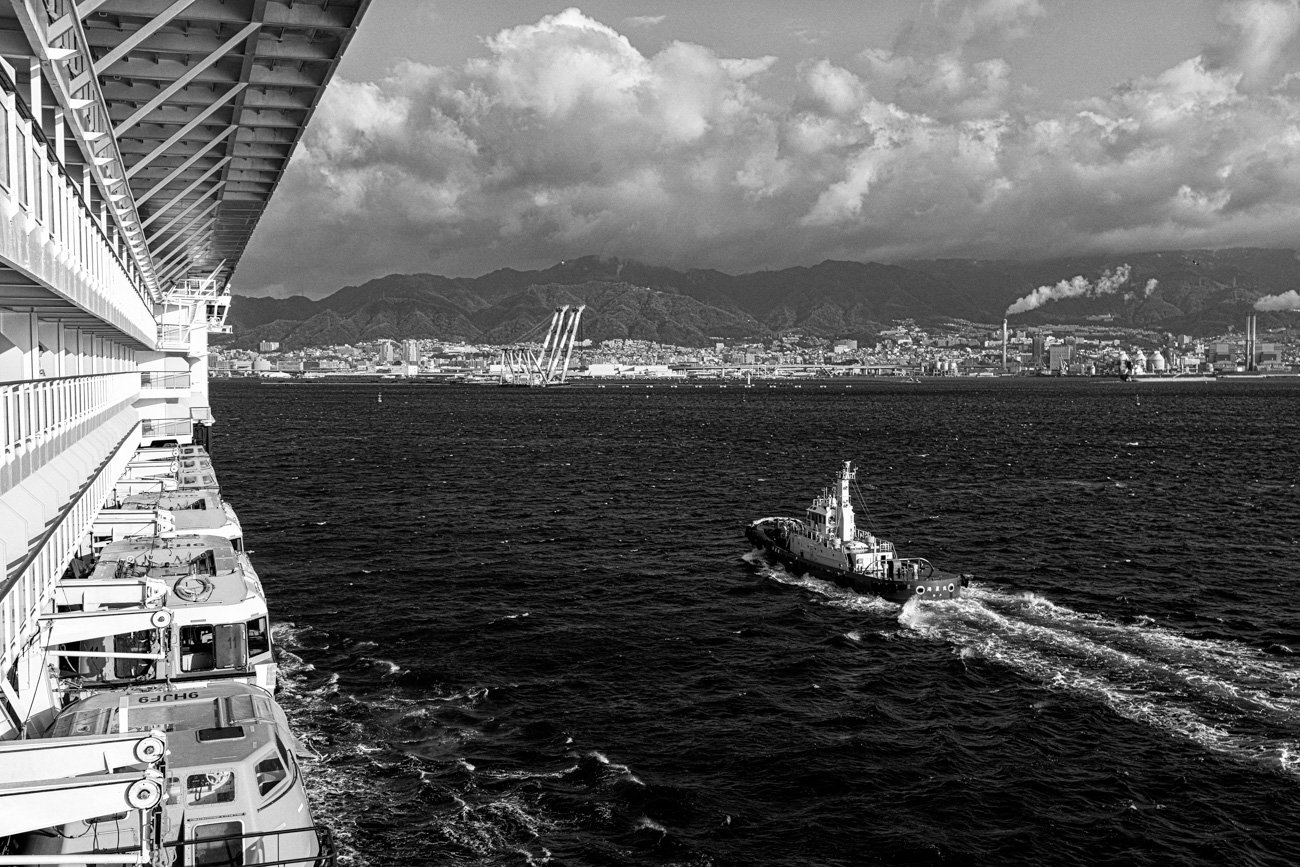 View from a cruise ship looking out over the ocean with a tugboat in the water, cityscape in the distance, and mountains under a cloudy sky in the background.