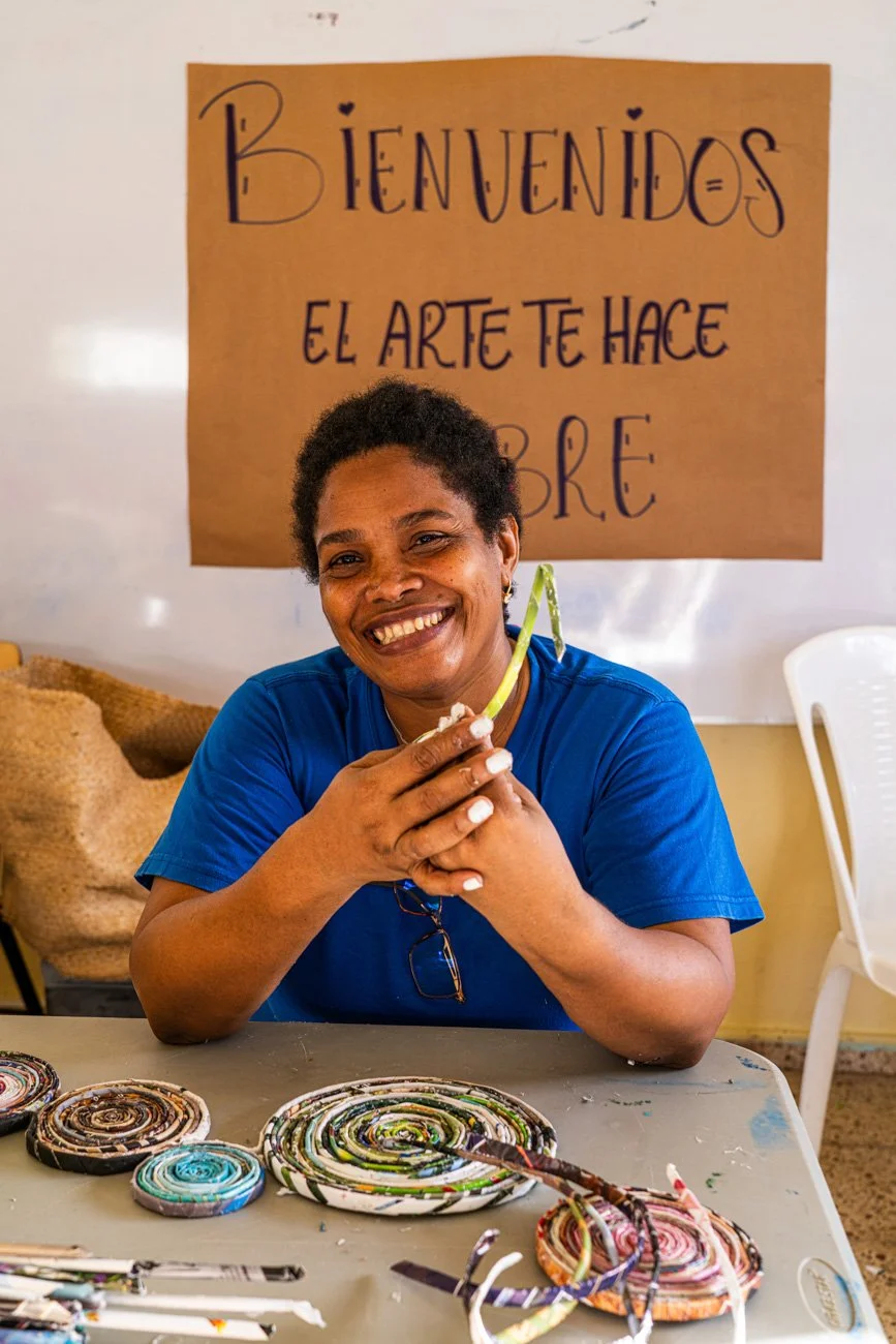 Smiling woman wearing a blue shirt sitting at a table with colorful handmade coiled art pieces, in front of a sign in Spanish that reads: "Bienvenidos. El arte te hace sentir libre."