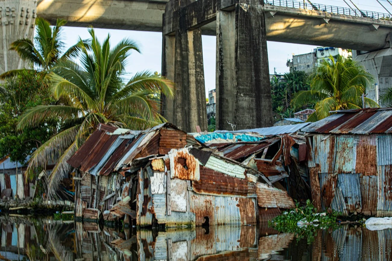 Dilapidated metal shacks in floodwaters beneath a concrete bridge with palm trees nearby.