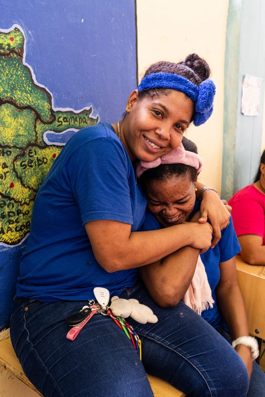 Two young women share a joyful hug, smiling and laughing. One is wearing a blue headband and a blue shirt, while the other has a pink headband and is also dressed in blue. They are sitting on a bench indoors with a colorful map of Guatemala on the wa
