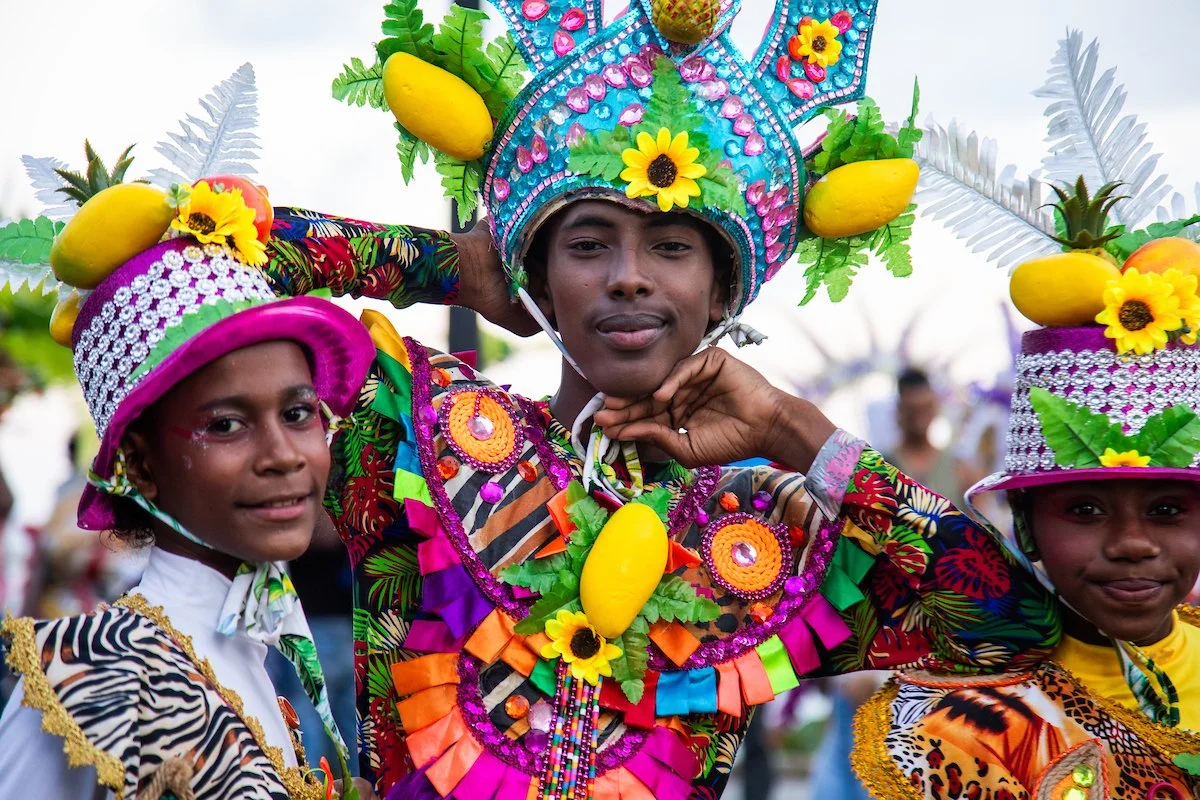 Three children dressed in colorful costumes with elaborate headdresses decorated with fruits, flowers, and beads, participating in a festive celebration or parade.