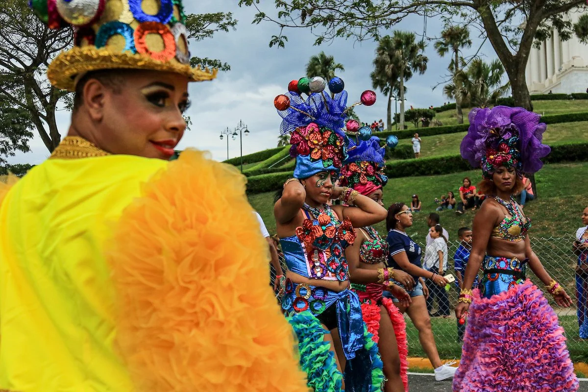 A woman in colorful carnival costume with a yellow ruffled sleeve and a large decorated hat, looking over her shoulder. Behind her, other women in vibrant costumes with elaborate headpieces are participating in a parade or festival in an outdoor park