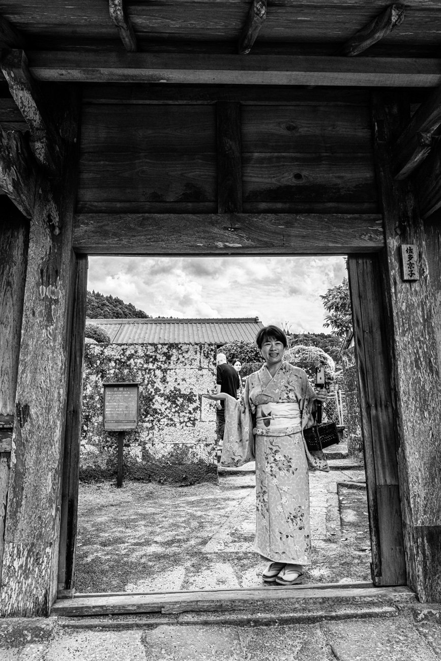 A woman in a traditional Japanese kimono standing at a wooden gate, smiling, with a garden and other people in the background.