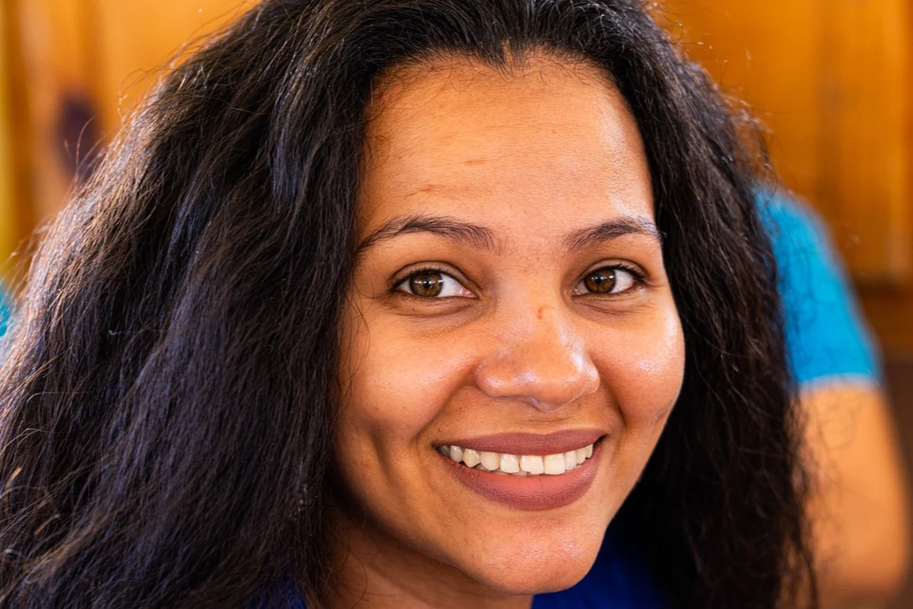 Close-up of a smiling woman with long dark hair and brown eyes, wearing a blue top, in a warm indoor setting.