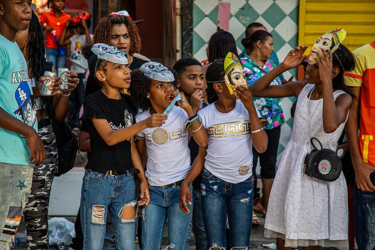 Children wearing masks and engaging in a street festival or celebration, with adults and colorful decor in the background.