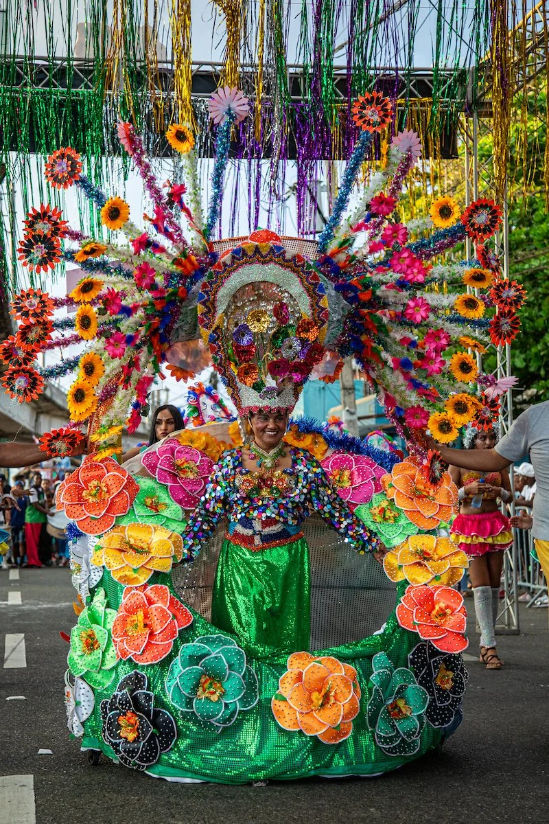 A person dressed in a vibrant floral costume with a large decorative headpiece featuring flowers, streamers, and colorful embellishments, participating in a parade celebration.