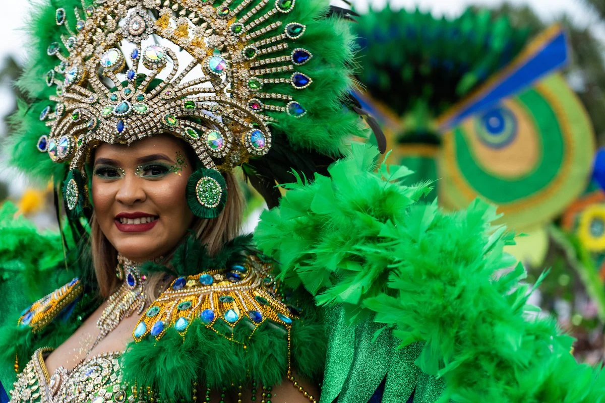 A woman dressed in a vibrant green carnival costume with elaborate beaded and rhinestone headpiece, feathered accessories, and bright makeup, smiling at a parade or festival.